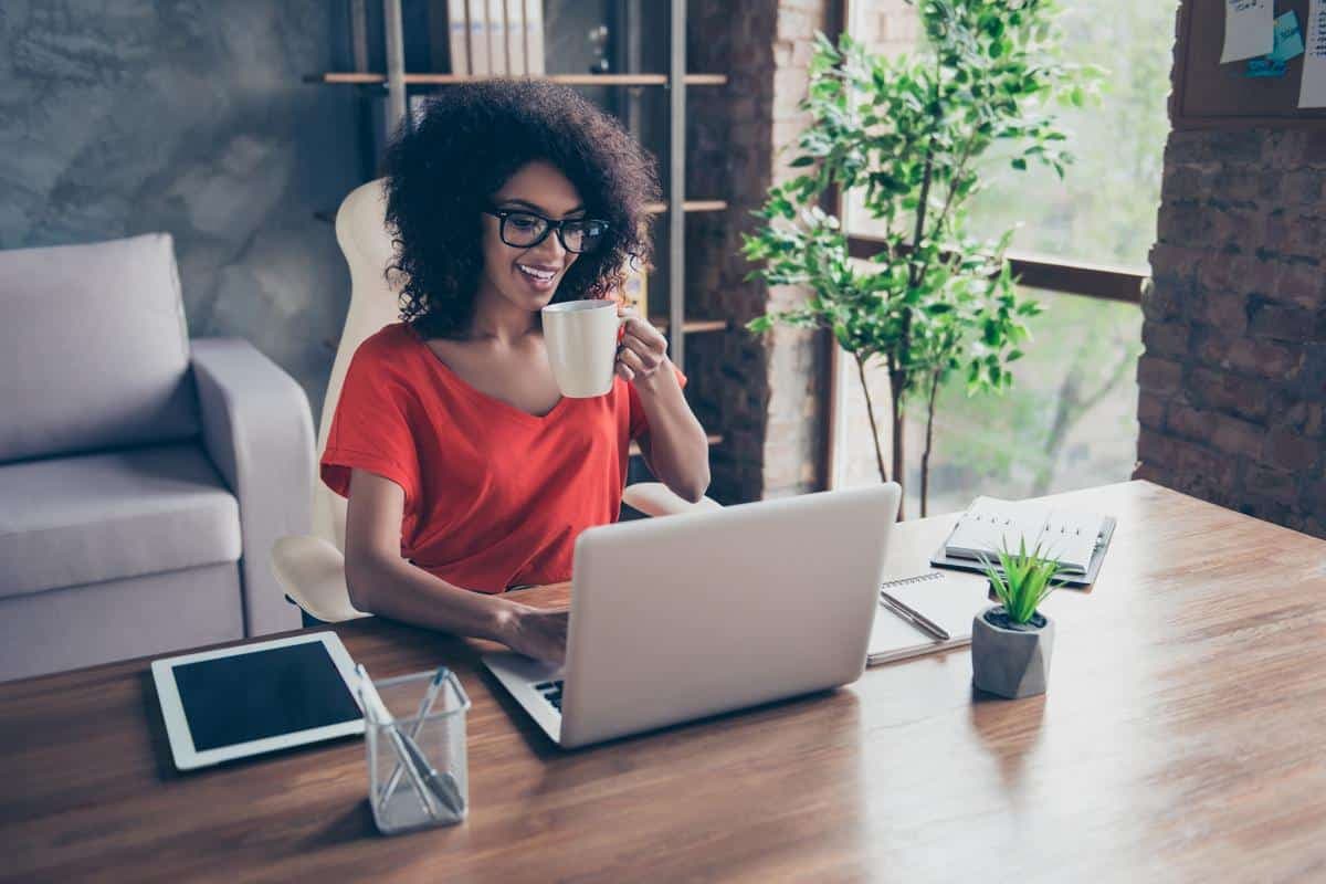 Portrait of cheerful positive freelancer sitting at desktop drinking hot beverage holding mug with tea typing on laptop chatting with friends enjoying free time using wi-fi internet