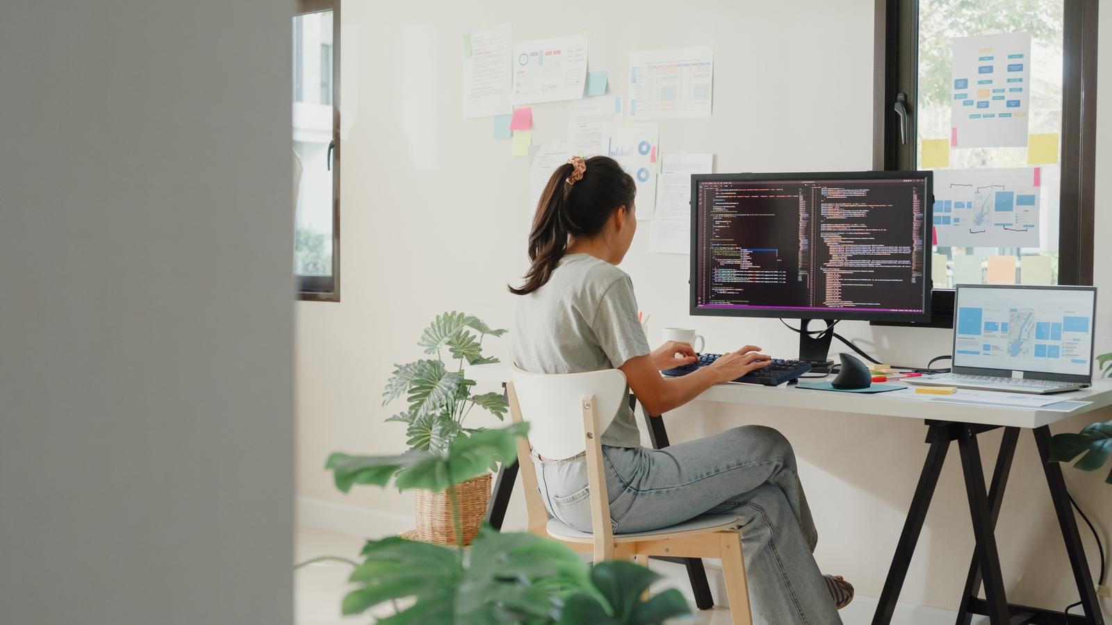 Side view of professional young Asia girl IT development programmer typing on keyboard coding programming fixing data code on computer screen and laptop on table in workroom at house office